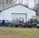 A collection of vintage car and truck bodies is scattered in front of a large white garage with a green metal roof. The scene is set on a grassy field with leafless trees in the background, indicating a late autumn or winter season. Various car parts, including an old blue car body and a gray truck, are visible. The vehicles appear to be in various stages of disrepair or restoration.